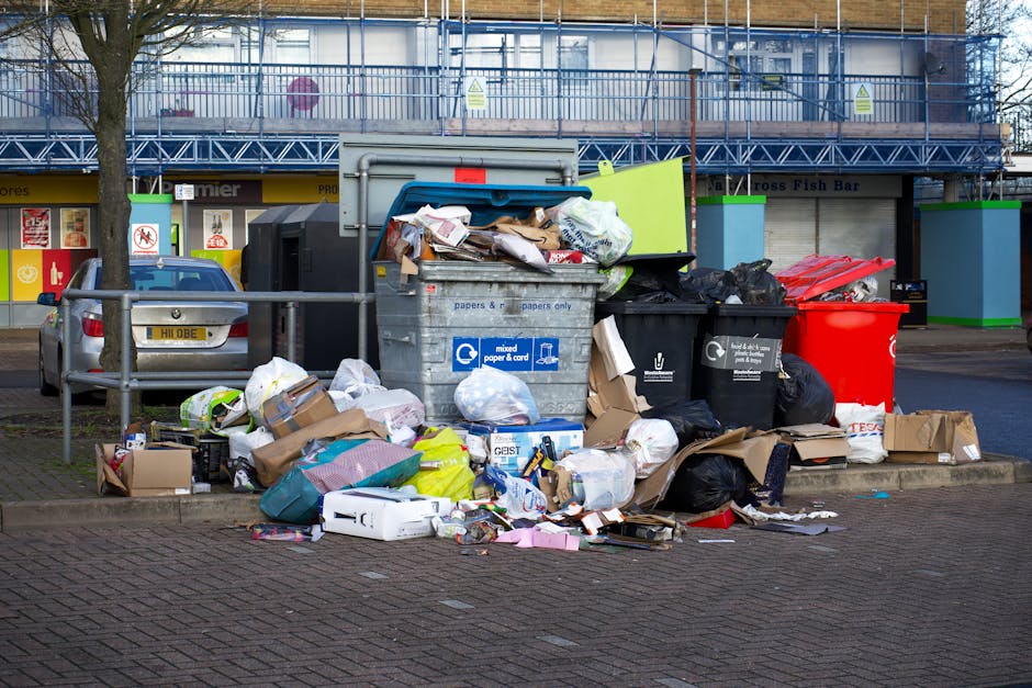 In the foreground, a large pile of mixed household waste and refuse bags is accumulated onto a paved sidewalk area, spilling over from a central metal recycling bin marked for paper and cardboard collection. The waste includes flattened cardboard boxes, crumpled paper, plastic bags, and discarded packaging materials in various colors such as brown, white, and plastic black sacks. Some items, like a white electronic device box and a green fabric bag, are partially open or crumpled, indicating recent disposal. To the left of the pile, a small cardboard box and additional paper waste rest on the ground. Behind the rubbish, a parked silver vehicle with a yellow license plate is visible, parked adjacent to a metal railing that separates the sidewalk from the road. In the background, a building under renovation features blue scaffolding and protective sheeting, with retail units below, including signs for fish and grocery shops. The scene suggests an instance of local waste accumulation, possibly for private rubbish collection, with waste removal services like Waste Removal Stockwell handling this bulk rubbish clearance. Lighting is natural and slightly overcast, casting diffuse light over the scene, emphasizing the disorder and variety of waste materials present.