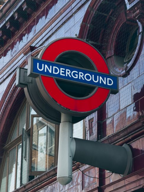 A close-up of a London Underground station sign featuring a circular red border with a horizontal blue bar across the center displaying the word 'UNDERGROUND' in white capital letters. The sign is mounted on a grey pole attached to an urban building facade with reflective glass windows and reddish-brown brick and tile exterior walls. The background shows the building's architecture with a mix of glass and tiled surfaces, some parts reflecting the sky and surrounding structures. The scene is taken during daylight with natural light illuminating the area, emphasizing the clean lines and vibrant colors of the signage. While primarily serving transportation, this image relates contextually to city waste management and the importance of organized urban infrastructure, which includes waste collection and disposal services often associated with busy public spaces and transportation hubs, such as those managed by waste removal companies like Waste Removal Stockwell.