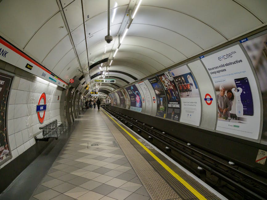 The image depicts a spacious underground railway station platform with a high, arched ceiling constructed of glass and metal, allowing natural light to illuminate the area. The platform features a smooth, dark surface with a tactile yellow safety strip along the edge closest to the train tracks. On the left side, there are a few passengers sitting on benches and waiting, dressed in casual clothing, with some individuals standing or walking along the platform. The station is lined with brick and stone archways that support the roof structure, and the overall environment appears clean and well-maintained. Visible train tracks run parallel to the platform, extending towards a dark tunnel at the far end of the station. The scene conveys a typical setting where passengers await a train, and the absence of litter or debris suggests a tidy environment, which could be relevant for private waste management or rubbish removal services operating within public transport facilities. Waste Removal Stockwell could assist in maintaining such cleanliness by providing professional rubbish clearance at similar transportation hubs, ensuring a safe and pleasant environment for travelers.